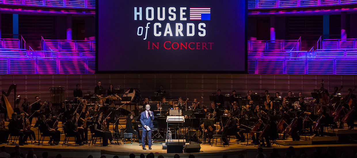 A man in a blue suit with a microphone is under the spotlight as a full orchestra sits down behind him with a House of Cards in Concert display in the back