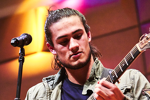 A man with long hair looks down as he plays the guitar behind a microphone