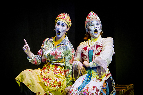 Two opera singers in bright colored clothes and white makeup sit on a bench as they sing in front of a black background
