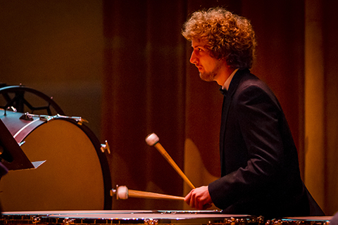 A man with curly hair and dark suit is playing the xylophone with 2 mallets he is holding in his hands.