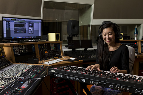 A woman wearing headphones plays the keyboard while sitting in a recording studio