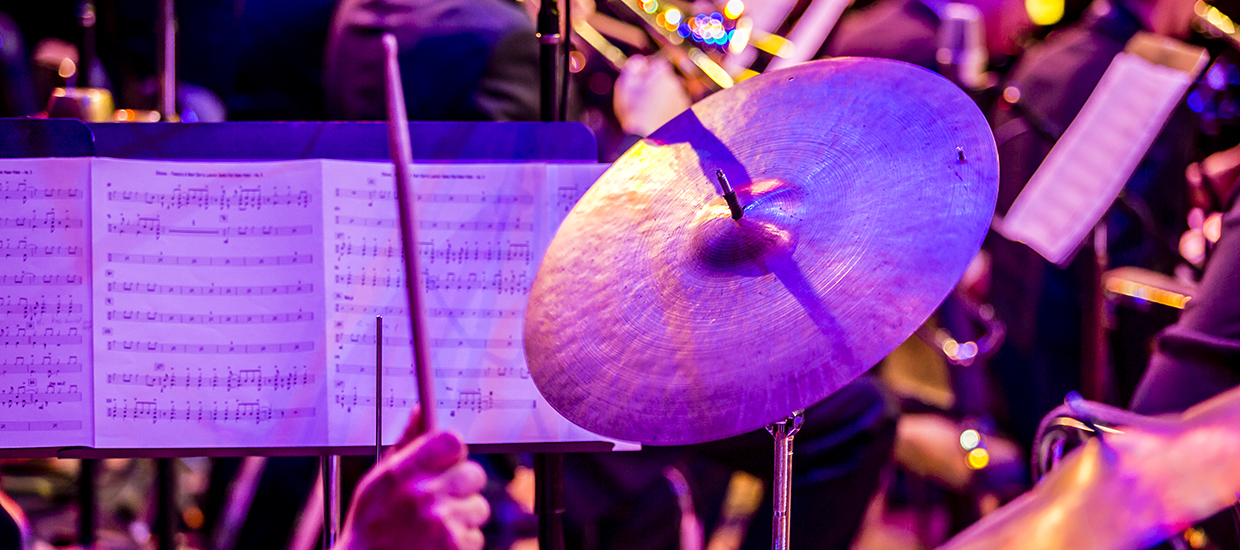 A drum stick heading towards a cymbal with sheet music being displayed in the background
