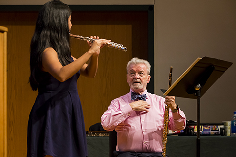 A student with long dark hair in a purple dress is playing the flute as a teacher looks on