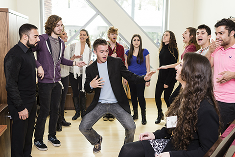 A group of students standing in a circle singing while a man in a suit jacket and jeans dances in the middle