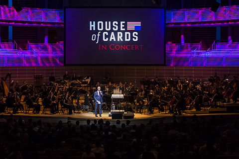 A man in a blue suit with a microphone is under the spotlight as a full orchestra sits down behind him with a House of Cards in Concert display in the back