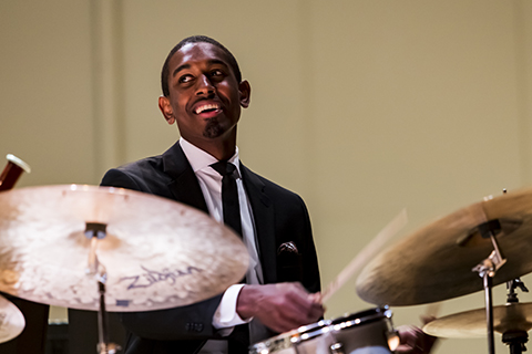 A drummer in a suit and tie smiles behind a drum-set as he looks off to his right.