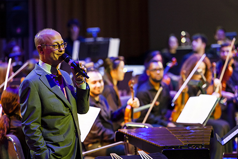 A man in a gray suit speaks into a microphone as an orchestra looks on behind him
